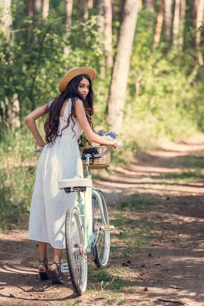 Young woman in straw hat and white dress walking with bicycle on trail in forest — Stock Photo