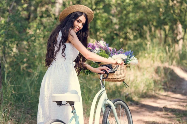 Hermosa mujer joven en sombrero de paja posando con bicicleta y flores en canasta de mimbre - foto de stock