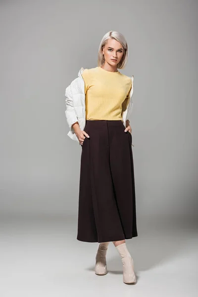 Серйозна молода жіноча модель в стильному осінньому вбранні позує руками в кишенях на сірому фоні — стокове фото
