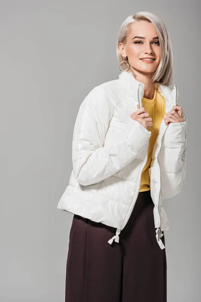 Усміхнена молода жінка в стильній білій куртці позує ізольовано на сірому фоні — стокове фото