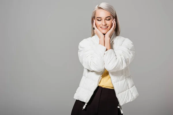Lächelnde Frau in weißer Jacke posiert mit Handflächen auf dem Gesicht isoliert auf grauem Hintergrund — Stockfoto