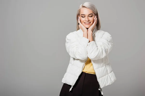 Mujer sonriente en chaqueta blanca posando con palmas de las manos en la cara aislada sobre fondo gris - foto de stock