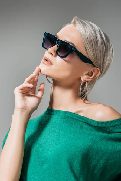 Stylische Frau mit Sonnenbrille und grünem Pullover posiert mit Hand am Kinn isoliert auf grauem Hintergrund — Stockfoto