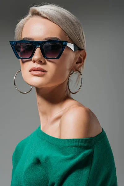 Modelo femenino atractivo en gafas de sol y suéter verde mirando a la cámara aislada sobre fondo gris - foto de stock