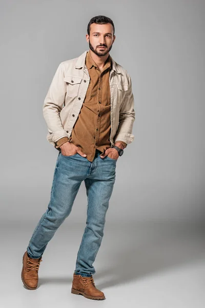 Красивый мужчина позирует в вельветовой рубашке и осенней куртке с руками в карманах джинсов, на сером — стоковое фото