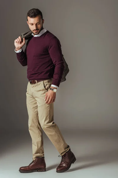Fashionable man posing in beige pants and burgundy sweater with autumn jacket on shoulder, on grey — Stock Photo