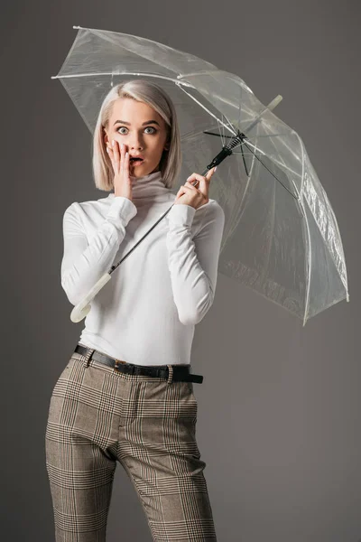 Elegant shocked girl in white turtleneck posing with fashionable transparent umbrella, isolated on grey — Stock Photo