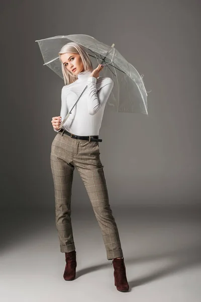 Приваблива блондинка в білому черепашку і сірі штани позують з прозорою парасолькою, на сірому — стокове фото