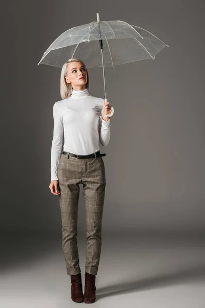 Elegante mujer de moda en cuello alto blanco y pantalones grises posando con paraguas transparente, sobre gris - foto de stock