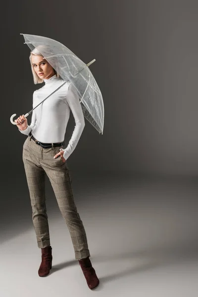 Menina elegante elegante em gola alta branca e calças cinza posando com guarda-chuva transparente, em cinza — Fotografia de Stock
