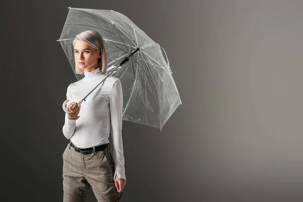 Menina atraente em gola alta branca posando com guarda-chuva transparente, isolado em cinza — Fotografia de Stock