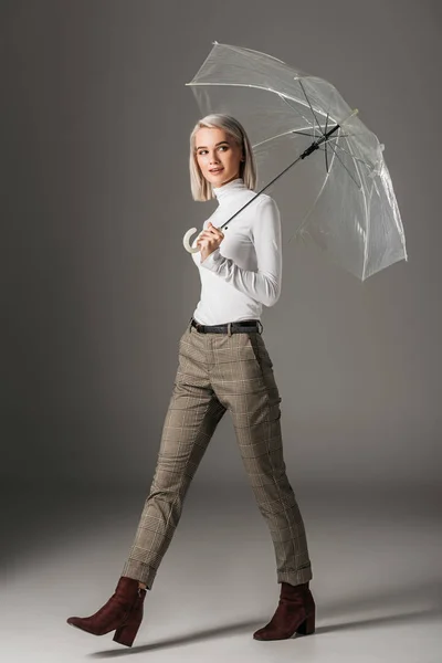 Elegante chica rubia en cuello alto blanco y pantalones grises caminando con paraguas transparente, en gris - foto de stock