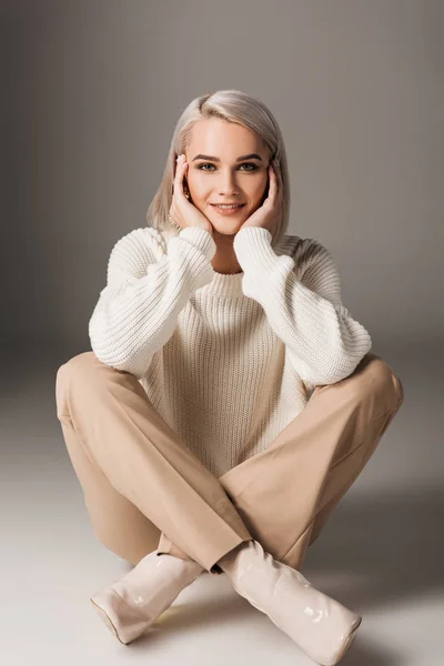 Schönes Mädchen sitzt in weißem Pullover und beiger Hose, auf grau — Stockfoto