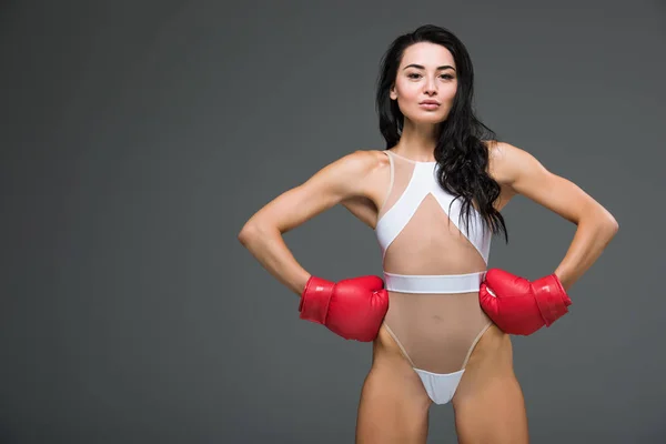 Seductive athletic woman in white leotard and boxing gloves standing with hands akimbo isolated on grey — Stock Photo