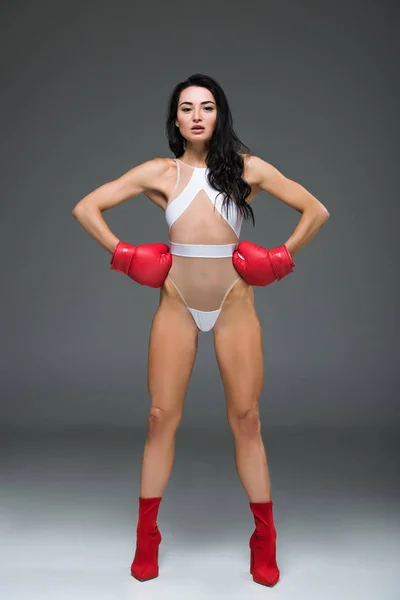 Mulher esportiva sexy em roupa de banho branca e luvas de boxe de pé com as mãos akimbo, em cinza — Fotografia de Stock