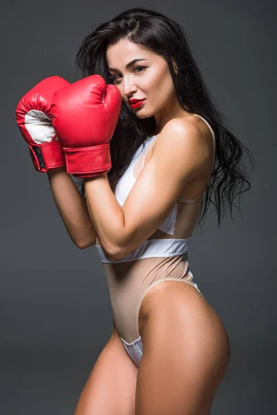 Вид сбоку на сексуальную спортивную женщину в белом платье и боксерских перчатках, смотрящую в камеру, изолированную от серого — стоковое фото
