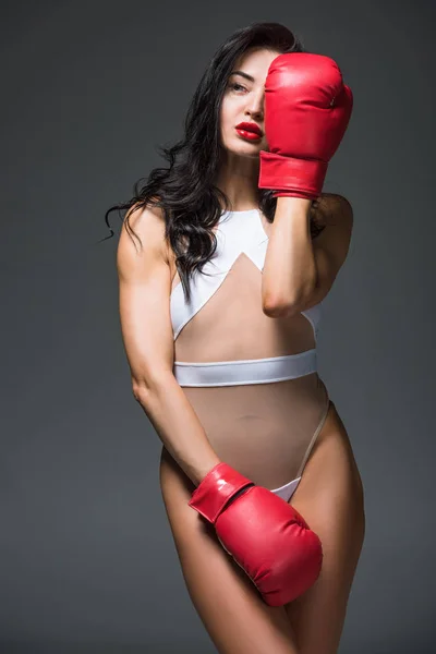 Sexy sportive woman in white swimwear covering eye with boxing glove isolated on grey — Stock Photo