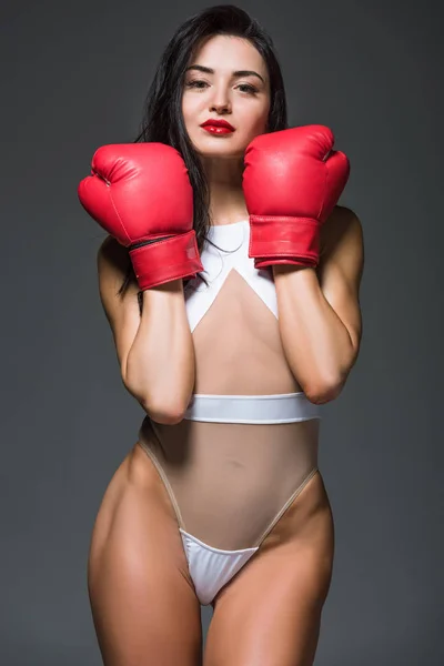 Sexy mujer deportiva en maillot blanco mostrando guantes de boxeo rojos aislados en gris - foto de stock