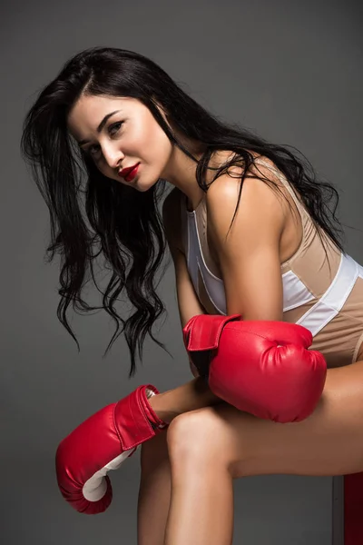 Вид збоку сексуальної спортивної жінки в білому купальнику і боксерських рукавичках, що сидять ізольовано на сірому — стокове фото