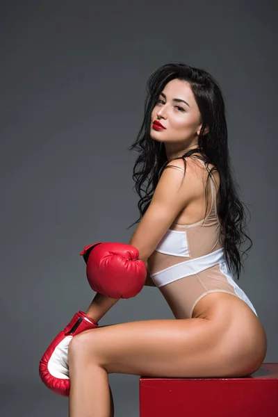 Side view of sexy sportive woman in white swimwear and boxing gloves sitting on red cube isolated on grey — Stock Photo