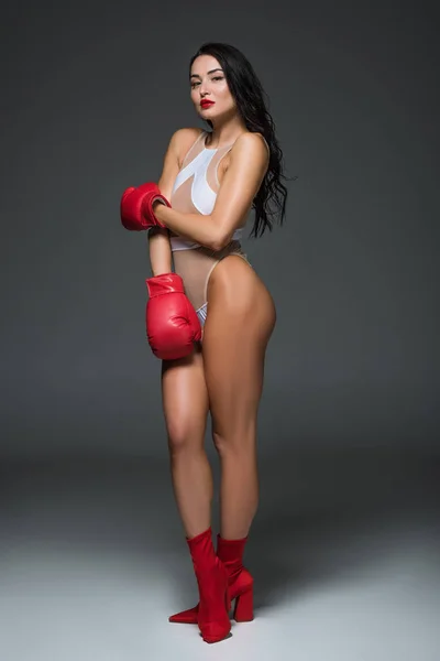 Sexy sportive woman in white leotard, red shoes and boxing gloves posing on grey — Stock Photo