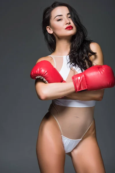 Atractiva mujer deportiva en maillot blanco y guantes de boxeo de pie con brazos cruzados aislados en gris - foto de stock