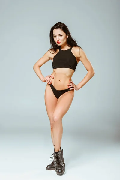 Сексуальна жінка в спортивному бюстгальтері і трусиках позує руками Акімбо, на сірому — стокове фото
