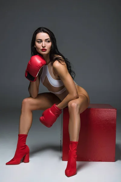 Sexy sportive woman in white swimwear and boxing gloves sitting on red cube on grey — Stock Photo