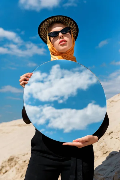 Fille attrayante à la mode posant avec miroir rond et reflet du ciel nuageux — Photo de stock