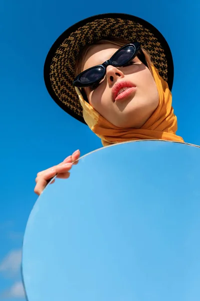Attrayant élégant fille posant dans des lunettes de soleil à la mode et tenant miroir rond avec réflexion sur le ciel bleu — Photo de stock