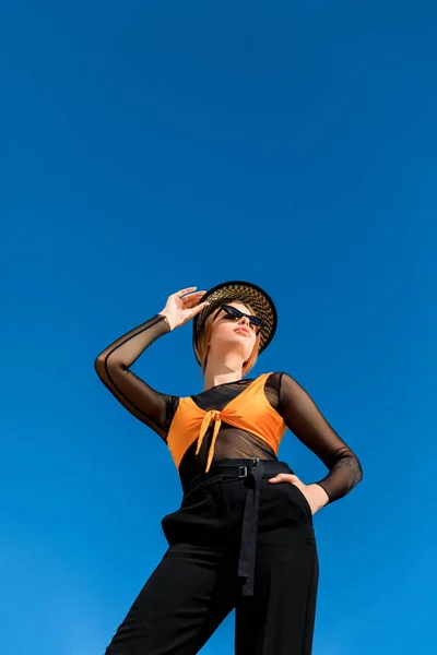 Vista inferior de la chica posando en gafas de sol de moda y sombrero elegante, cielo azul en el fondo - foto de stock
