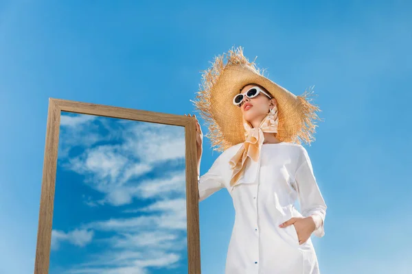 Mujer elegante posando en ropa blanca de moda, bufanda y sombrero de paja cerca del espejo con el reflejo del cielo azul - foto de stock