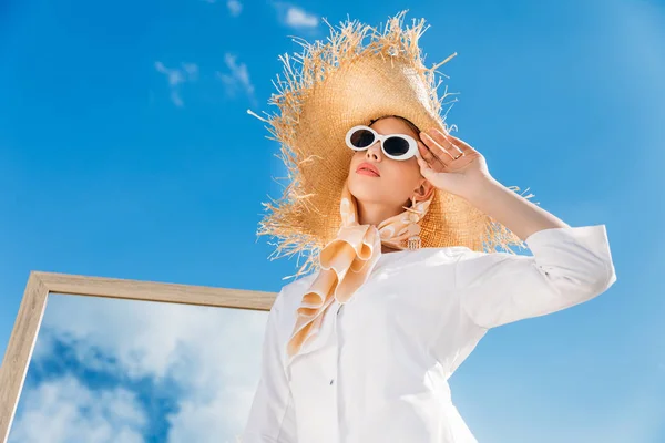 Menina elegante posando em óculos de sol, roupas brancas, cachecol e chapéu de palha perto do espelho no dia ensolarado — Fotografia de Stock