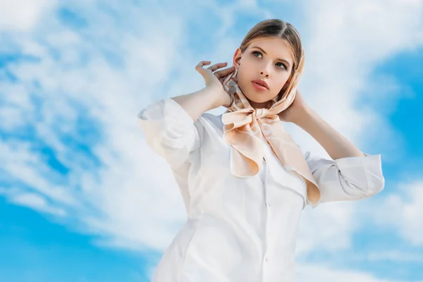 Вид знизу красивої стильної жінки в білому одязі і шовковому шарфі, з блакитним хмарним небом — стокове фото