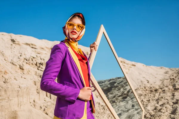 Beau modèle élégant en vêtements à la mode posant près du miroir sur une dune de sable — Photo de stock