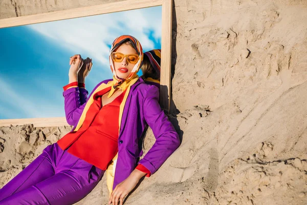 Mulher na moda atraente em roupas elegantes deitado na areia e espelho com reflexão sobre o céu — Fotografia de Stock