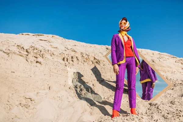 Bellissimo modello in abiti colorati in posa vicino specchio sulla duna di sabbia con cielo blu — Foto stock