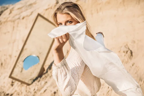 Stylish woman posing in white clothes with silk scarf near mirror on sand — Stock Photo