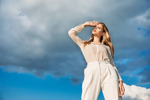 Vista inferior de mujer rubia elegante posando en ropa blanca con cielo nublado en el fondo - foto de stock