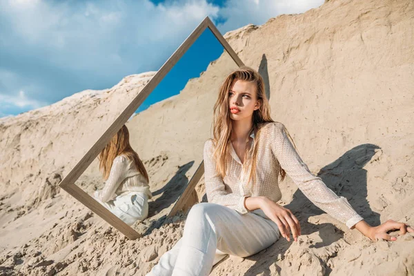 Елегантна молода жінка позує в білому одязі біля дзеркала на піску — стокове фото