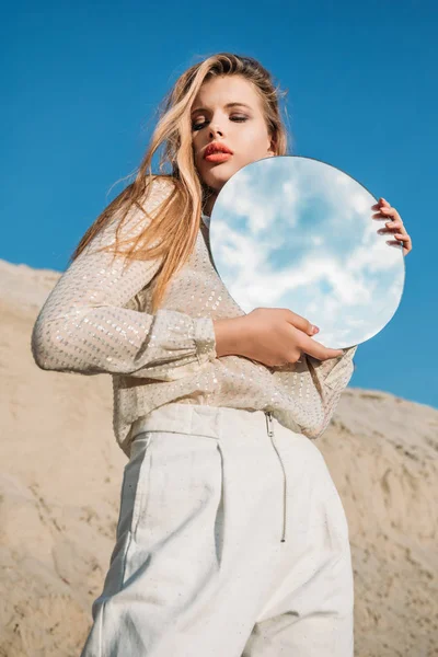 Modello biondo alla moda in vestiti bianchi che tengono lo specchio rotondo con riflessione di cielo nuvoloso — Foto stock
