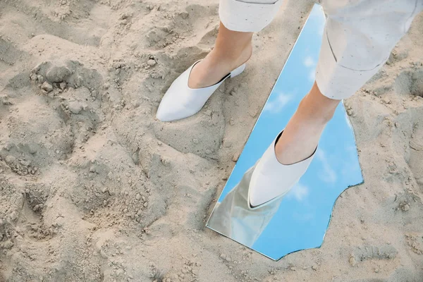 Visão parcial do modelo em pé na areia e pedaço de espelho com reflexão do céu azul — Fotografia de Stock