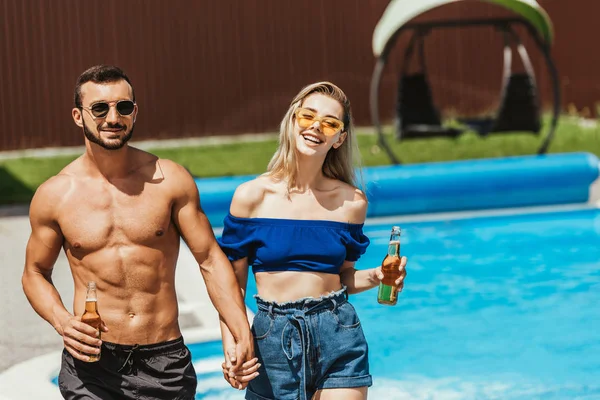 Casal com garrafas de cerveja de mãos dadas à beira da piscina — Fotografia de Stock
