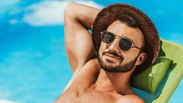 Bel homme en lunettes de soleil et chapeau de paille bronzer sur un transat au bord de la piscine — Photo de stock