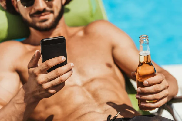 Vista recortada del hombre usando un teléfono inteligente y sosteniendo la botella de cerveza - foto de stock