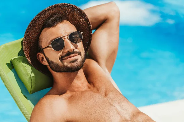 Homem bonito em óculos de sol e chapéu de palha relaxante na espreguiçadeira na piscina — Fotografia de Stock