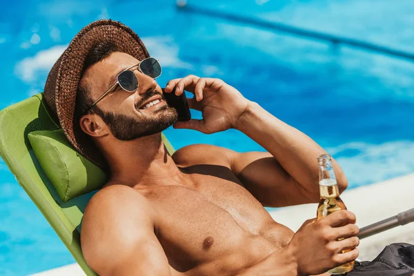 Hombre en gafas de sol hablando en el teléfono inteligente y sosteniendo la botella de cerveza en la cama al lado de la piscina - foto de stock