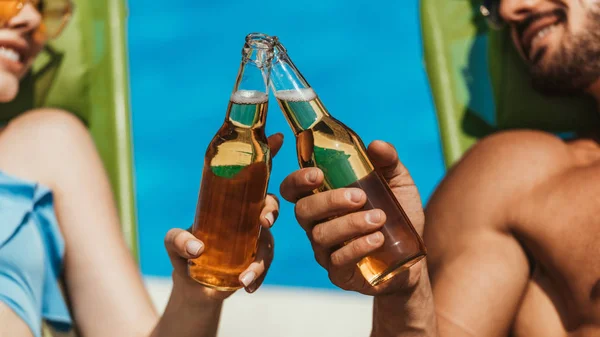 Vista recortada de pareja tintineo con botellas de cerveza en tumbonas - foto de stock