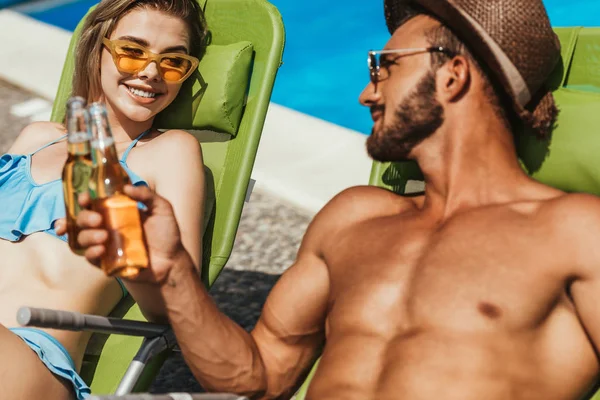 Paar klingelt mit Bierflaschen, während es sich am Pool auf Sonnenliegen ausruht — Stockfoto