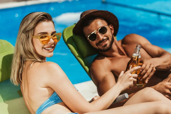 Coppia sorridente in occhiali da sole clinking con bottiglie di birra mentre sdraiato su lettini — Foto stock